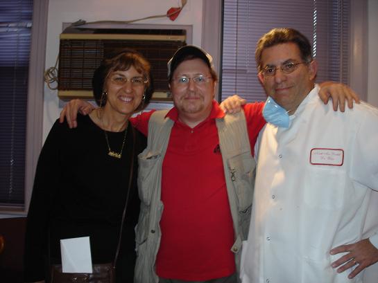 In the image, there are three individuals posing for a photograph  two adults standing on either side of another adult who appears to be seated. The central figure is wearing a white lab coat and glasses, suggesting they might be a scientist or medical professional. The person in the middle is smiling and seems to be in a jovial mood, while the other two individuals are also smiling. They all appear to be in an indoor setting with a window visible in the background.