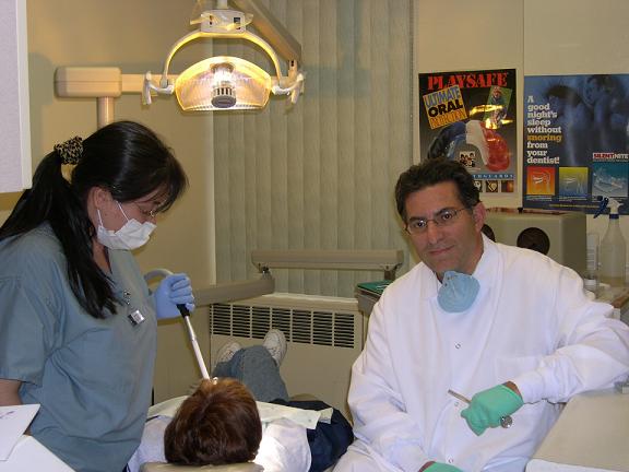 In the image, a man in a white lab coat with a stethoscope around his neck is standing next to a woman wearing scrubs, both are in a room that appears to be a dental office based on the presence of dental equipment and a patient lying down.