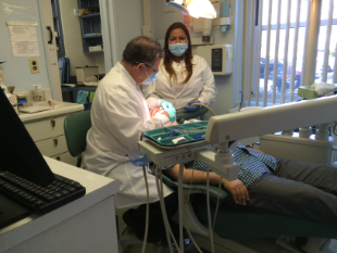 An image depicts three individuals in a dental office setting, with one person receiving dental care while another appears to be assisting and the third person observes.