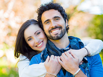 A man and woman embrace each other with smiles, both wearing casual clothing and standing outdoors against a blurred background.