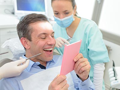 The image shows a man sitting in a dental chair with a smile on his face, holding up a pink card, while a dentist, wearing gloves and a mask, stands behind him, smiling and looking at the card.