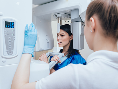 A woman wearing blue gloves stands next to a large white scanner machine while another person looks on with interest.